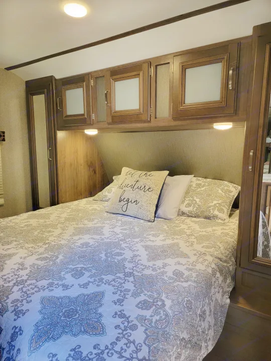 Cozy RV rental bedroom featuring a patterned bed, overhead wood cabinets, and integrated bedside lighting for travel adventures.. Keystone RV Passport Grand Touring 2018