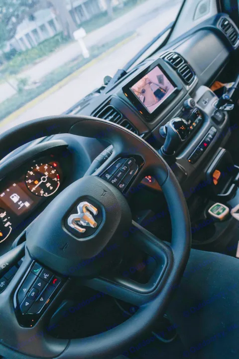 Inside a modern RV rental van: steering wheel with Ram logo, dashboard, and infotainment screen displaying a camera view.. Ram ProMaster 3500 2024