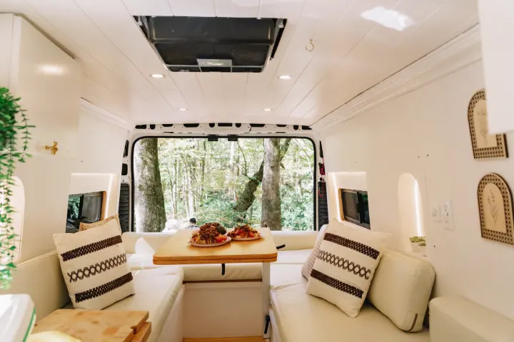 Inside this stylish rv rental camper, a dining area with a meal overlooks a serene forest, offering a cozy retreat.. Ram ProMaster 3500 2024