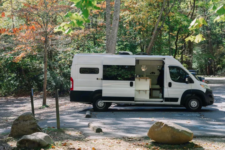 White camper van (RV rental) parked in an autumn forest with its side door open, revealing a cozy interior for a nature getaway.. Ram ProMaster 3500 2024
