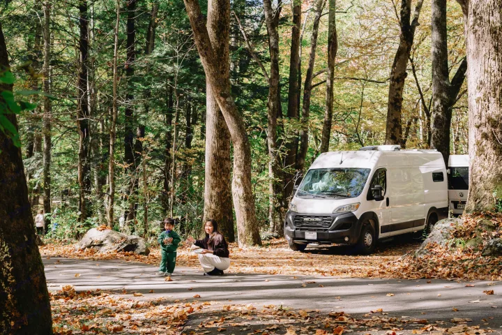 A family enjoys an autumn day with their RV rental parked in a serene forest, surrounded by vibrant fall foliage.. Ram ProMaster 3500 2024