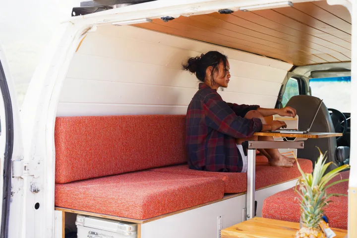 Woman remote working on laptop in RV rental camper van, featuring a custom wood interior and cozy orange cushions.. Ford Econolline 250s 2005