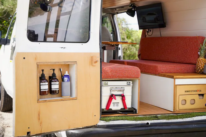 This rv rental shows a camper van interior with custom door storage for toiletries, a cozy red-orange bench, table, TV, and a Nomad Kitchen.. Ford Econolline 250s 2005