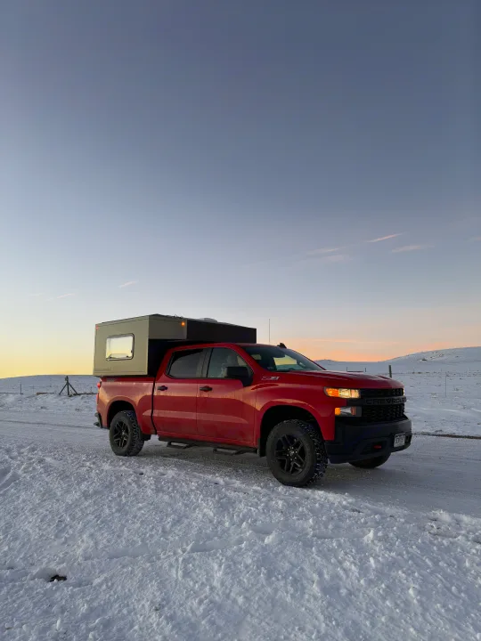 A red Chevy pickup truck with a gray truck camper on a snowy road at sunset. Explore winter landscapes with this RV rental.. Chevrolet Silverado 1500 Trail Boss 2020