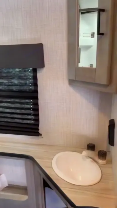 Compact RV rental bathroom featuring a sink, wood-look vanity, mirror cabinet, and window blind.. Heartland RVs Mallard 2021