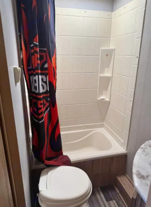 Compact RV rental bathroom with a small tub, shower, and toilet. Features a black and red patterned shower curtain.. Coachmen Freedom Select Express 28.7 2019