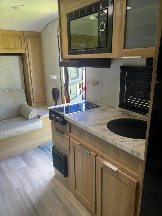 This rv rental interior features a modern kitchen with a microwave, stovetop, sink, and light wood cabinetry, plus a cozy couch.. Forest River Salem FSX 2023