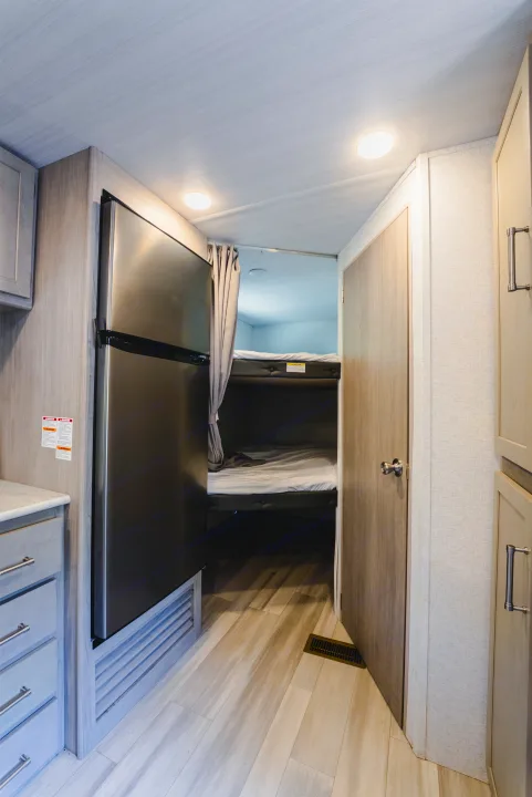 An RV rental's light-toned interior, featuring a black refrigerator, wood cabinetry, and bunk beds with a gray privacy curtain.. Keystone Springdale 2024