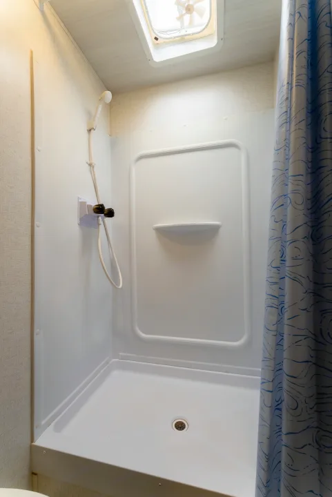 Clean, compact RV rental shower stall with white walls, handheld showerhead, faucet, vent fan, and blue patterned curtain.. Keystone Springdale 2024