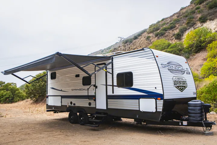 An RV rental: a white Springdale travel trailer with its awning extended, parked on a dirt lot against a green hillside.. Keystone Springdale 2024