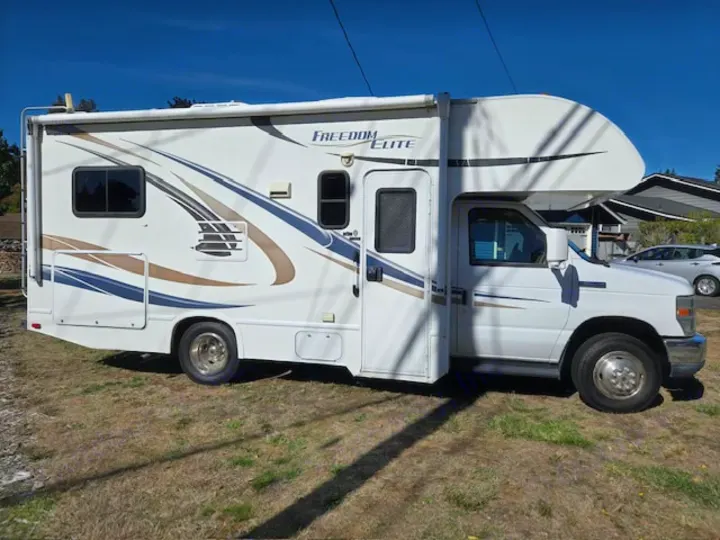 Side view of a white "Freedom Elite" Class C rv rental with blue and brown decals, parked on grass on a sunny day.. Thor Freedom Elite 2015