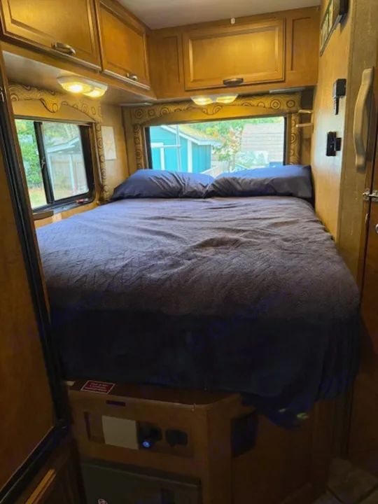 This RV rental bedroom features a large dark blue bed, wooden cabinets, and windows showing outdoor scenery for a comfortable stay.. Thor Freedom Elite 2015