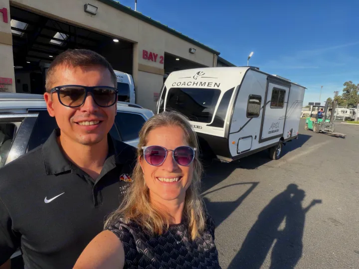 A happy couple poses with their white Coachmen Apex travel trailer, an RV rental, outside a service bay.. Coachmen Apex Nano 194BHS 2026