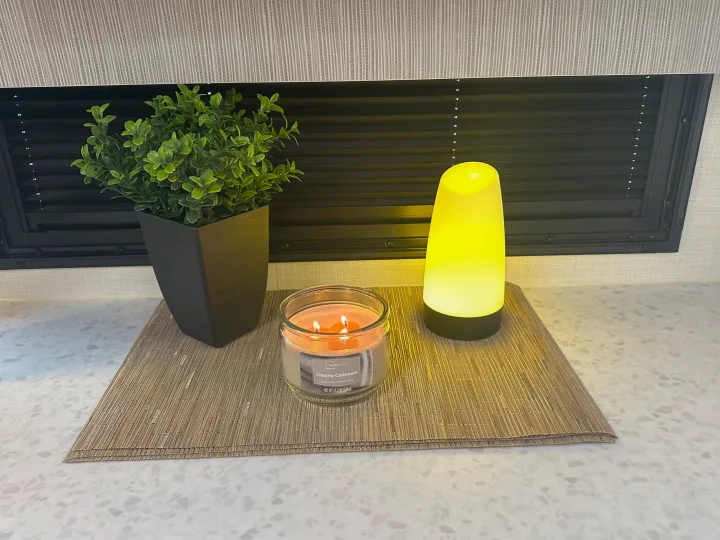 Cozy RV rental interior: a lit Creamy Cashmere candle, green plant, and warm yellow lamp on a speckled countertop with blinds.. Forest River Tracer Ultra Lite 2025
