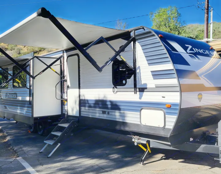 This white and blue Zinger travel trailer RV rental, with its awning extended and entry steps down, is ready for camping.. Crossroads RV Zinger 2021