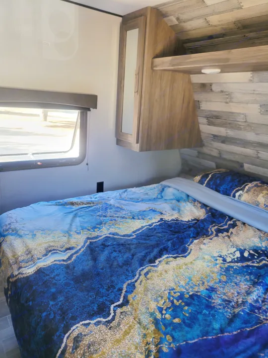 Inside an RV rental, this cozy bedroom features a bed with vibrant blue & gold bedding, a window, and wood cabinetry.. Crossroads RV Zinger 2021