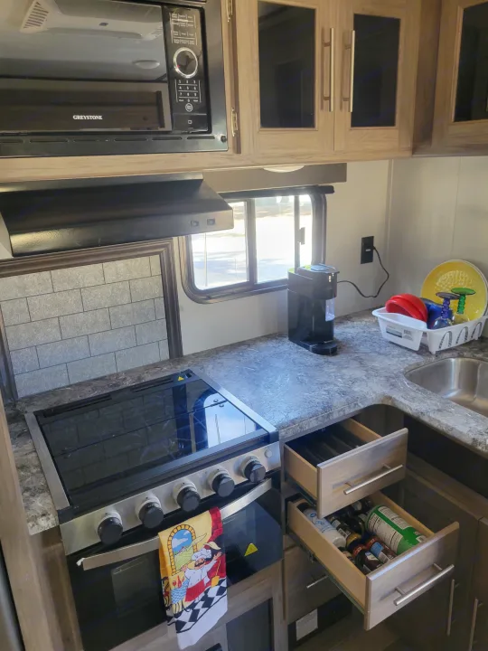 Compact RV rental kitchen featuring a Greystone microwave, gas range, sink, coffee maker, and open drawers with spices.. Crossroads RV Zinger 2021