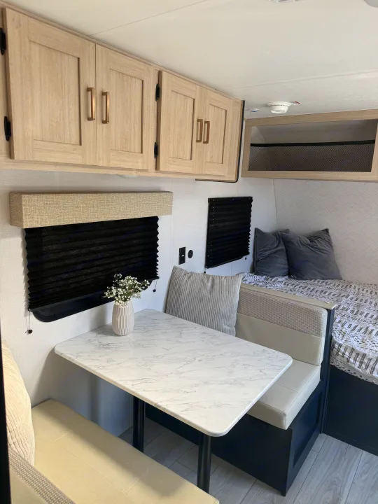 Light and airy rv rental interior featuring a dinette with a marble-look table, overhead cabinets, and a cozy bed with pillows.. Forest River Avenger LT 2025