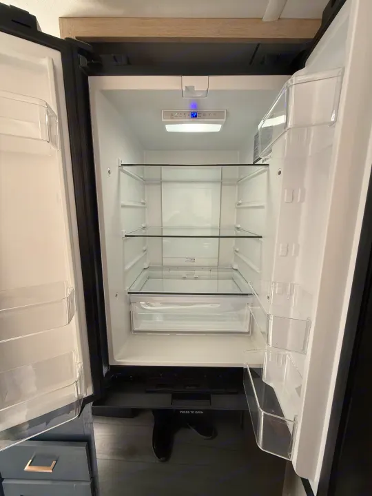 Inside an open, empty RV rental refrigerator, featuring clear glass shelves, plastic door bins, and a digital control panel.. Forest River Avenger LT 2025