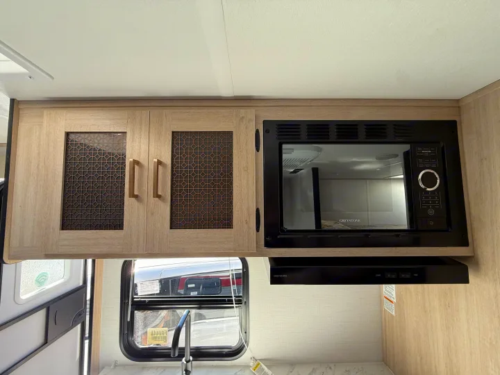Inside an RV rental kitchen: light wood cabinets, Greystone microwave, range hood, sink, and window view.. Forest River Avenger LT 2025
