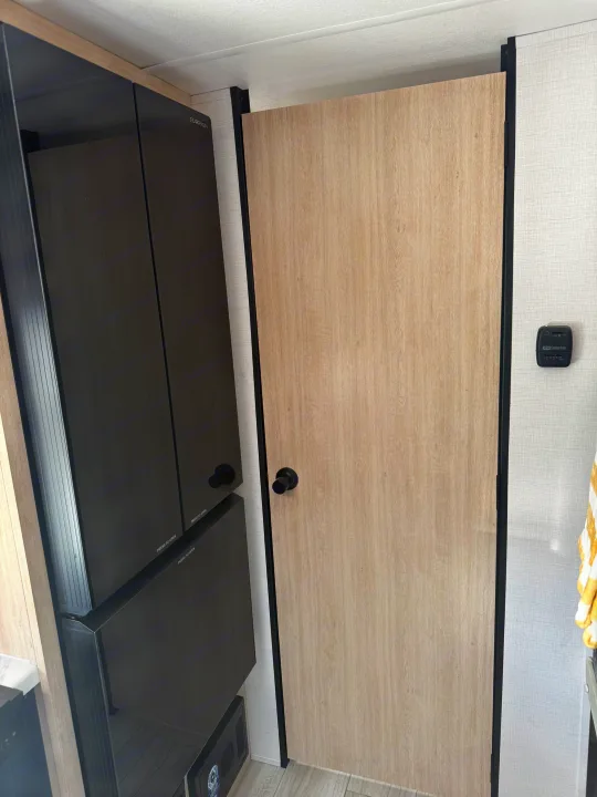 RV rental interior with a light wood door, a dark Furrion refrigerator unit, and a Suburban thermostat on the textured wall.. Forest River Avenger LT 2025