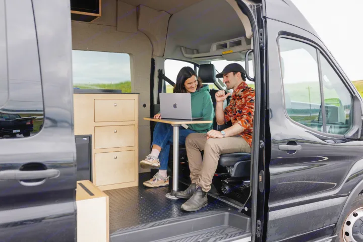 Smiling couple using a laptop inside an RV rental's cozy, converted workspace, ideal for digital nomad remote work on the road.. Ford Transit 2023