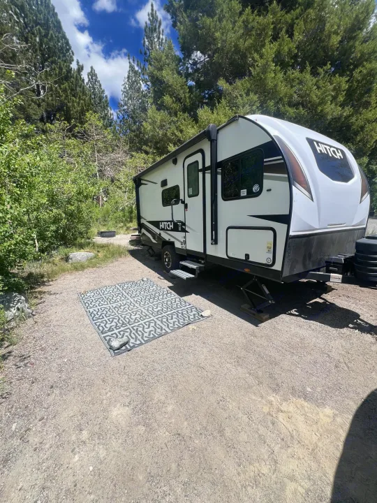 A white RV rental travel trailer with an outdoor mat is parked on a gravel campsite, surrounded by tall trees under a blue sky.. Cruiser RV Hitch 2024