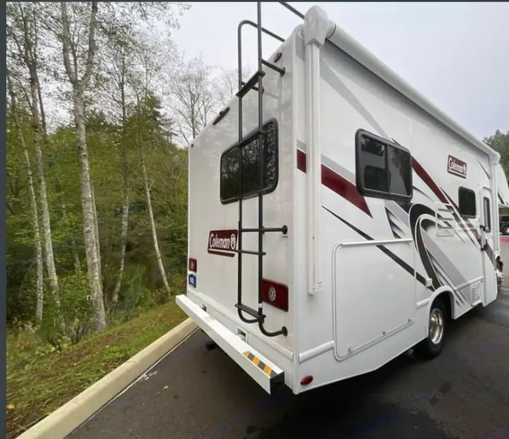 Explore the outdoors with this Coleman RV rental. A white motorhome with burgundy stripes, ladder, and awning, parked by a forest.. Thor Freedom Elite 2026