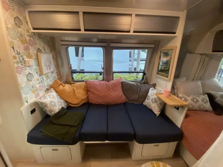 Cozy rv rental interior with a blue cushioned bed/seating, floral wallpaper, and colorful pillows, creating a welcoming travel space.. Airstream Bambi 2024