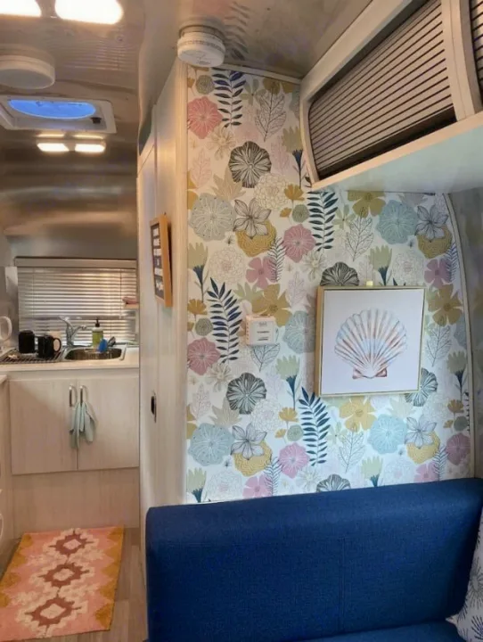 Stylish RV rental interior: floral wallpaper, blue couch, kitchen sink, and seashell art create a vibrant, cozy space.. Airstream Bambi 2024