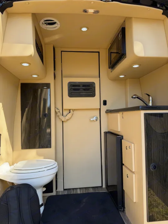 Modern RV rental interior: beige cabinetry, dark wood accents. Features compact kitchen with sink, microwave, fridge, and toilet.. Ultimate Toys Ultimate Coach 2024
