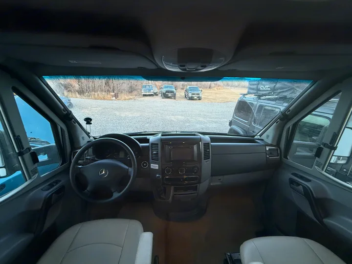 Inside this RV rental, the driver's seat view of a Mercedes-Benz Sprinter's dashboard and steering wheel, with trucks visible outside.. Winnebago View 2014