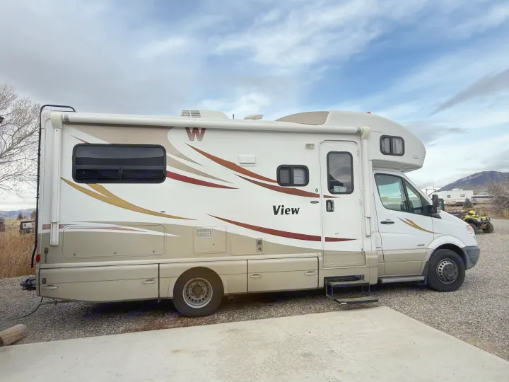 Side view of a parked Winnebago View Class C motorhome (rv rental) in white and tan, with steps extended on gravel.. Winnebago View 2014