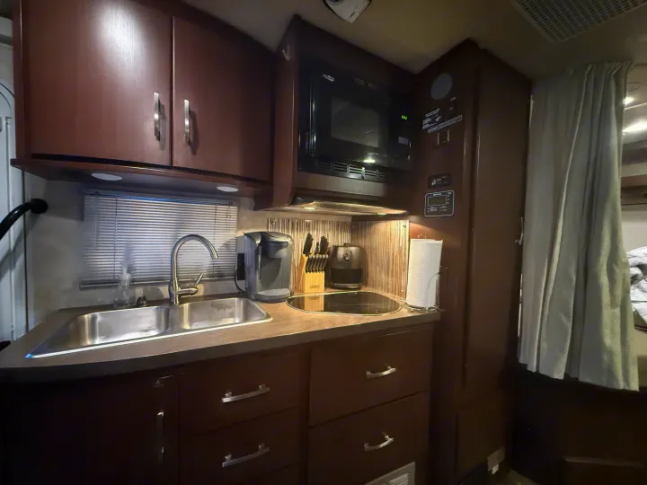 Experience comfort in this RV rental kitchen, featuring dark wood cabinets, a double sink, microwave, Keurig, and cooktop.. Winnebago View 2014