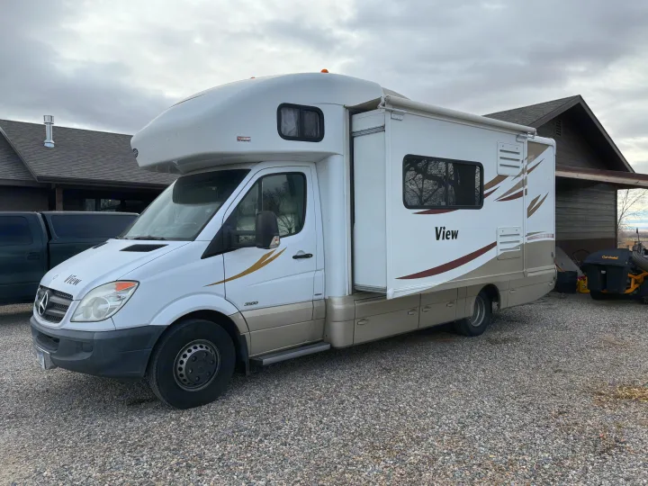 White & tan Winnebago View RV rental on gravel, slide-out extended, parked by a house. Ideal for travel.. Winnebago View 2014