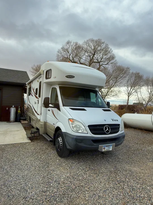 A white Mercedes-Benz Sprinter rv rental, branded 'View', is parked on gravel under an overcast sky.. Winnebago View 2014