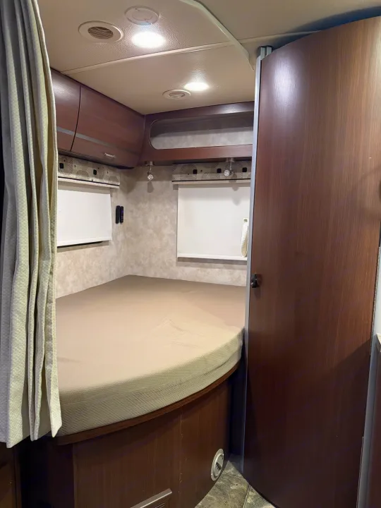 Cozy RV rental bedroom featuring a curved bed, overhead storage, window shades, and a privacy door, ideal for travel.. Winnebago View 2014