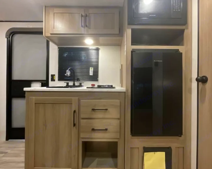 Compact RV rental kitchen features a sink, stove, microwave, and Furrion refrigerator, with light wood cabinetry and storage.. Keystone RV Coleman 2025