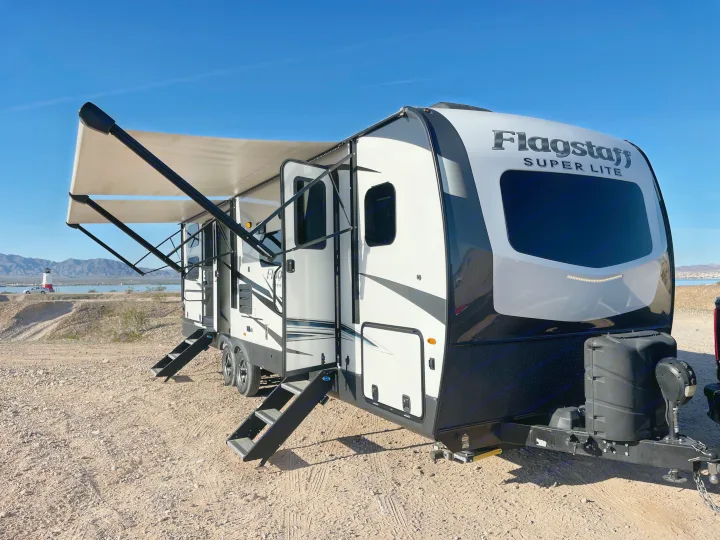 Flagstaff Super Lite RV rental: a white and black travel trailer with awning extended, parked by a lake and mountains.. Forest River Flagstaff Super Lite 2021