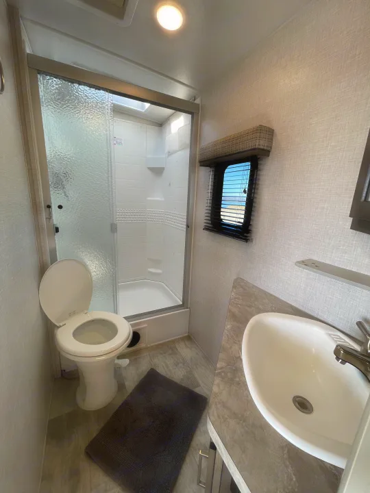 Clean rv rental bathroom featuring a white toilet, glass shower, sink on a gray counter, and a window with scenic views.. Forest River Flagstaff Super Lite 2021
