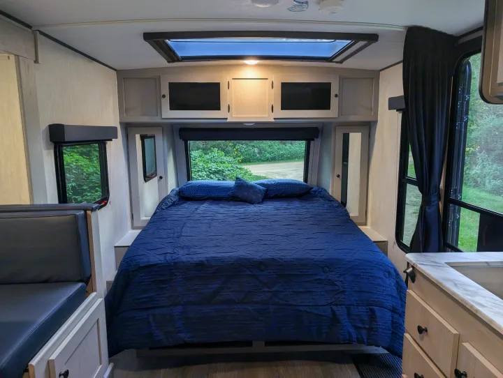 Modern rv rental interior with a comfy blue bed, skylight, dinette, and outdoor views of greenery.. Coachmen Apex Nano 2025
