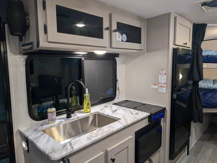 Modern RV rental kitchen featuring a sleek sink, induction cooktop, microwave, and fridge, with bunk beds nearby.. Coachmen Apex Nano 2025