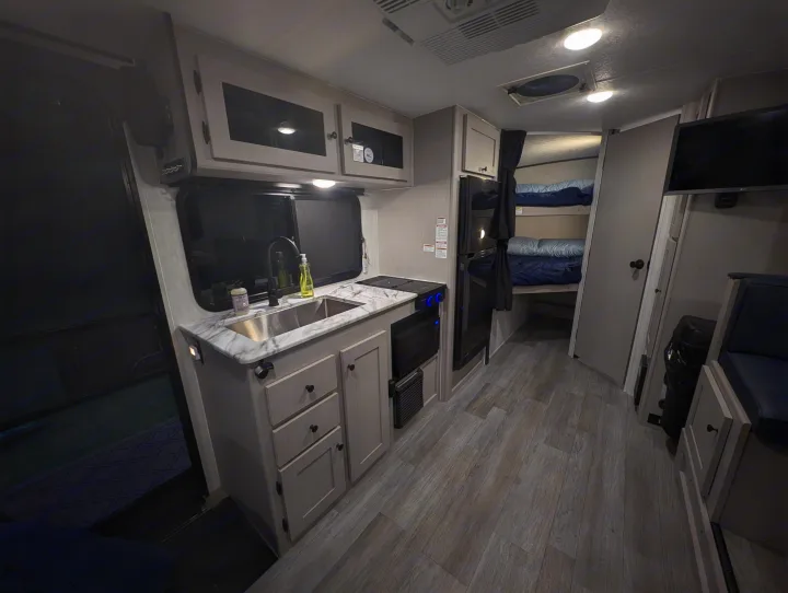 Cozy RV rental interior with a modern kitchen, sink, stovetop, microwave, and bunk beds. Features light wood-look flooring.. Coachmen Apex Nano 2025