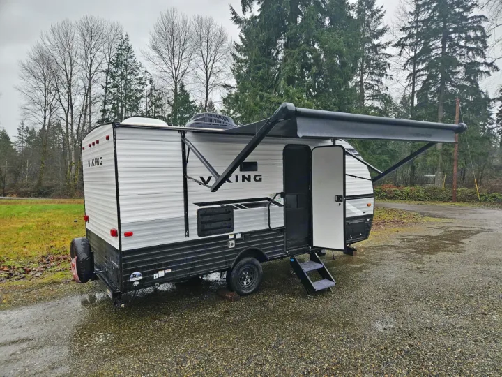 RV rental travel trailer: A white and black unit with an extended awning, parked on gravel next to trees under a cloudy sky.. Forest River Viking 4K 2023