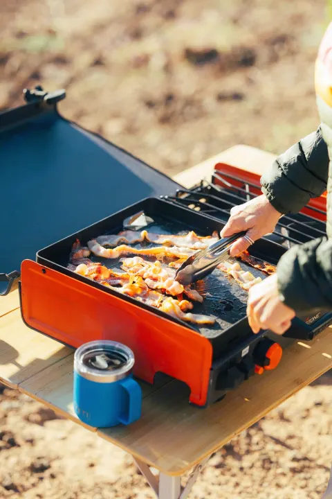 Outdoor breakfast with your RV rental! A person cooks bacon on a portable griddle, with a blue Yeti mug on a folding table.. Toyota Tundra 2019