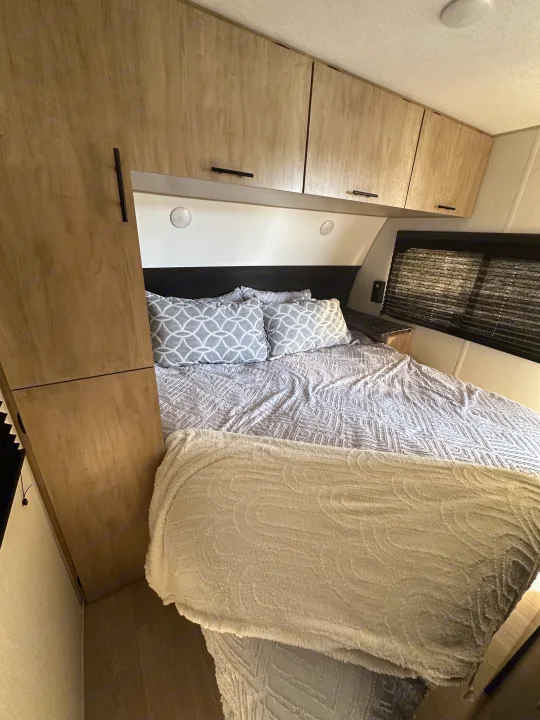Cozy RV rental bedroom with patterned bed linens, a fuzzy blanket, overhead wood cabinets, and window.. Forest River Cherokee Grey Wolf 2026