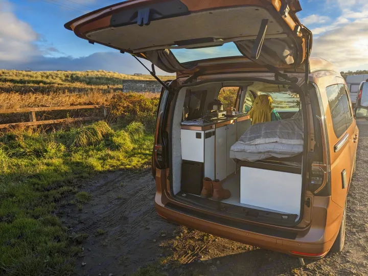 RV rental campervan interior, featuring a kitchen & bed, revealed through its open tailgate by grassy dunes.. Volkswagen Caddy Maxi 2023