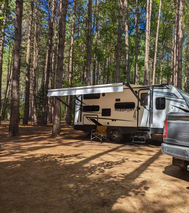 A white Passport Sport RV rental is parked in a sunny pine forest campsite with its awning extended. A truck is attached.. Keystone RV Passport SL 2021