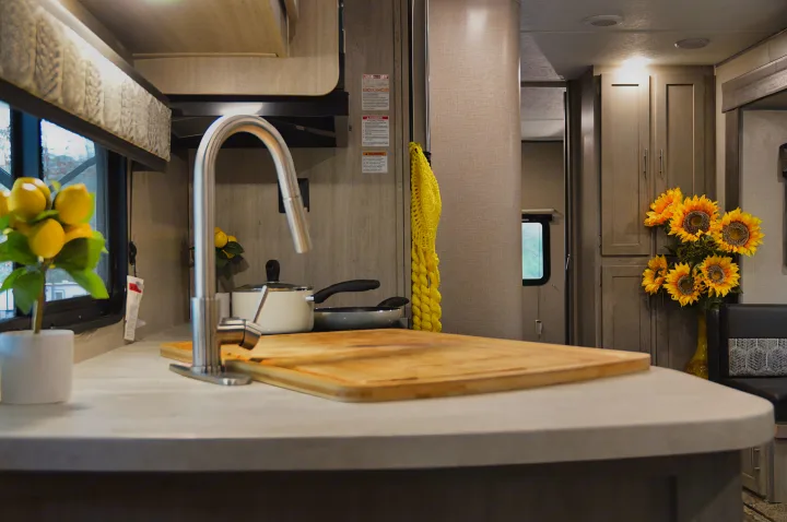 Bright RV rental kitchen with a modern stainless steel faucet, wooden cutting board, and cheerful sunflowers. Cabinets and window visible.. Coachmen Catalina 2022