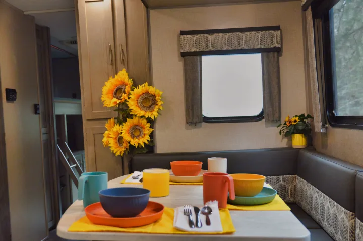 RV rental interior with a bright dining area. Colorful dishes on yellow placemats, sunflowers, and a glimpse of bunk beds.. Coachmen Catalina 2022